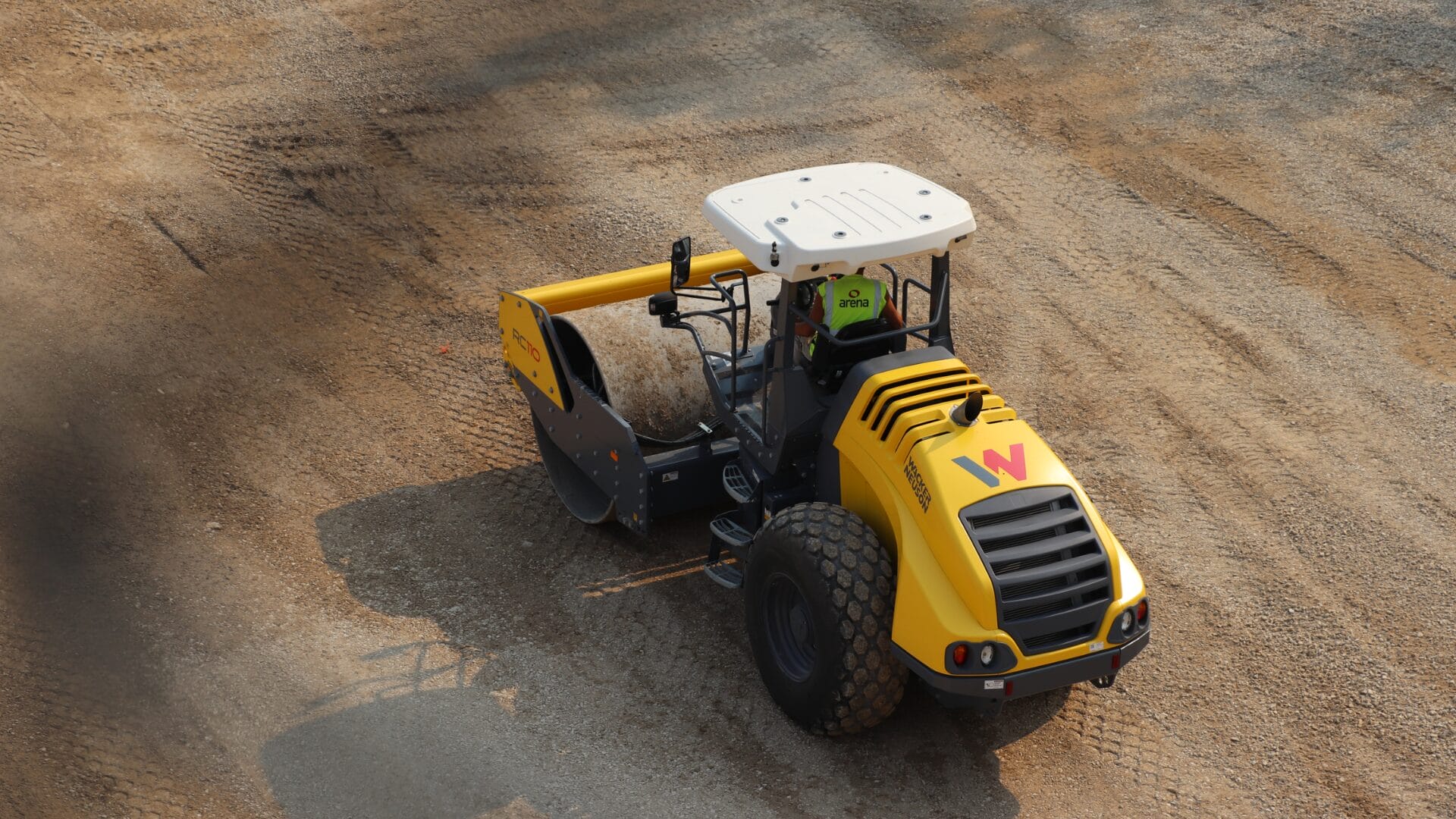 A yellow road roller compactor with a white roof rests on a gravel surface. Its large drum, vital for choosing the right roller for soil compaction, is clearly visible. The ground bears tracks from previous construction activity, showcasing its role in landscaping or road work.