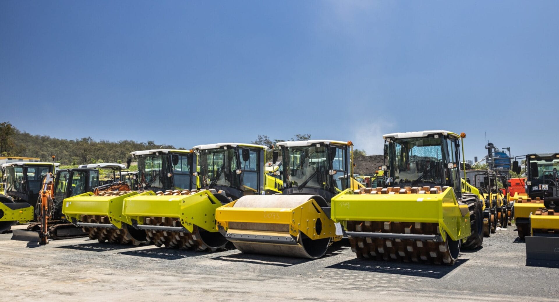 A row of bright yellow heavy equipment, including steamrollers and construction vehicles, is parked on a dirt lot under a clear blue sky. Trees are visible in the background, and the area provides safe storage at what appears to be a construction site or machinery storage area.