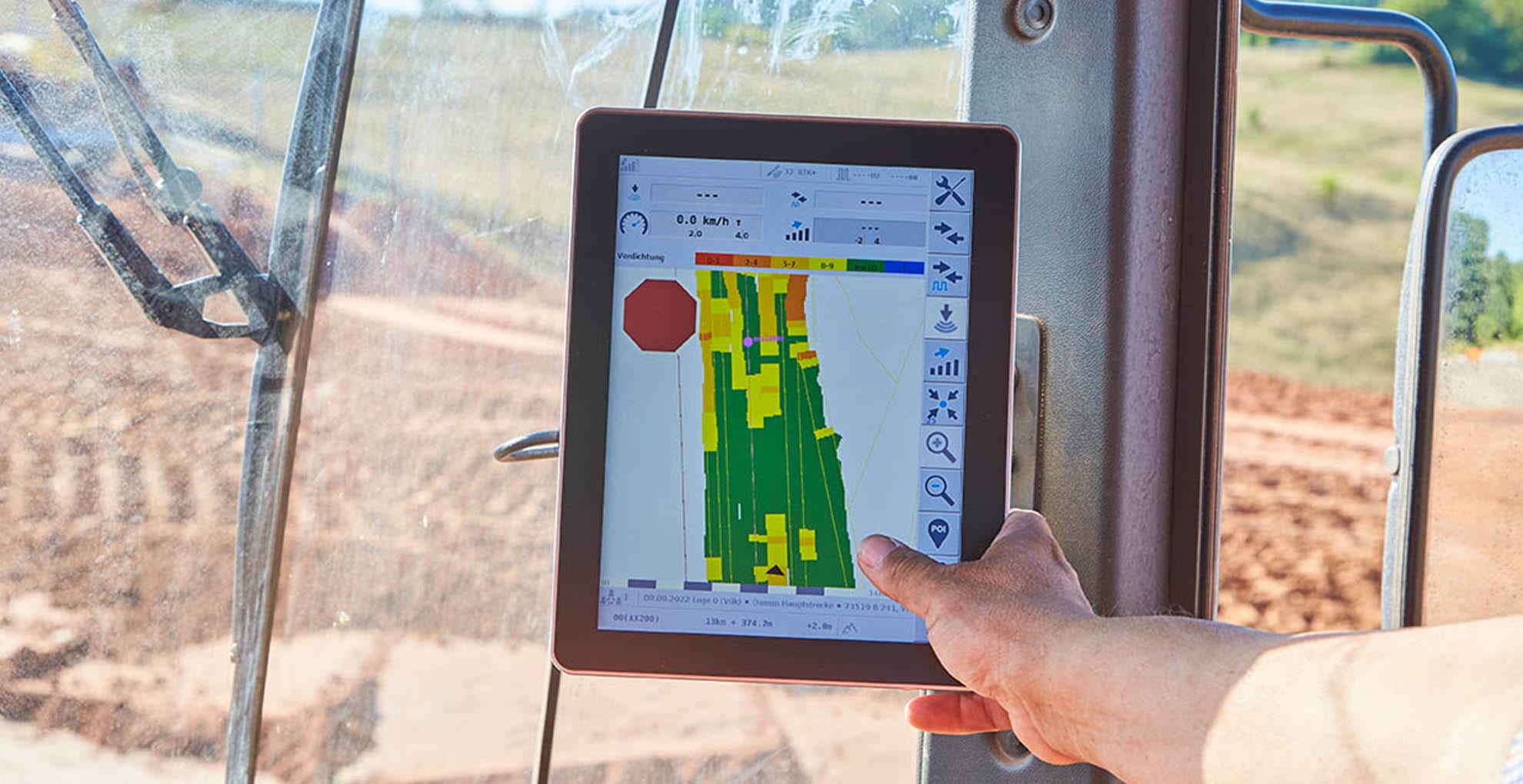 A person's hand holds a tablet displaying a colorful map and various data metrics inside a vehicle. The scene outside the vehicle shows a dirt and grassy landscape, suggesting the equipment might be used for agricultural or construction purposes, showcasing advanced technology in action.