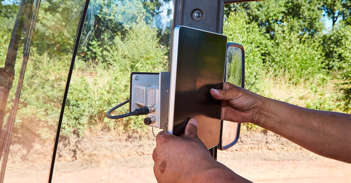 Hands holding a tablet near construction equipment inside a vehicle with trees outside.