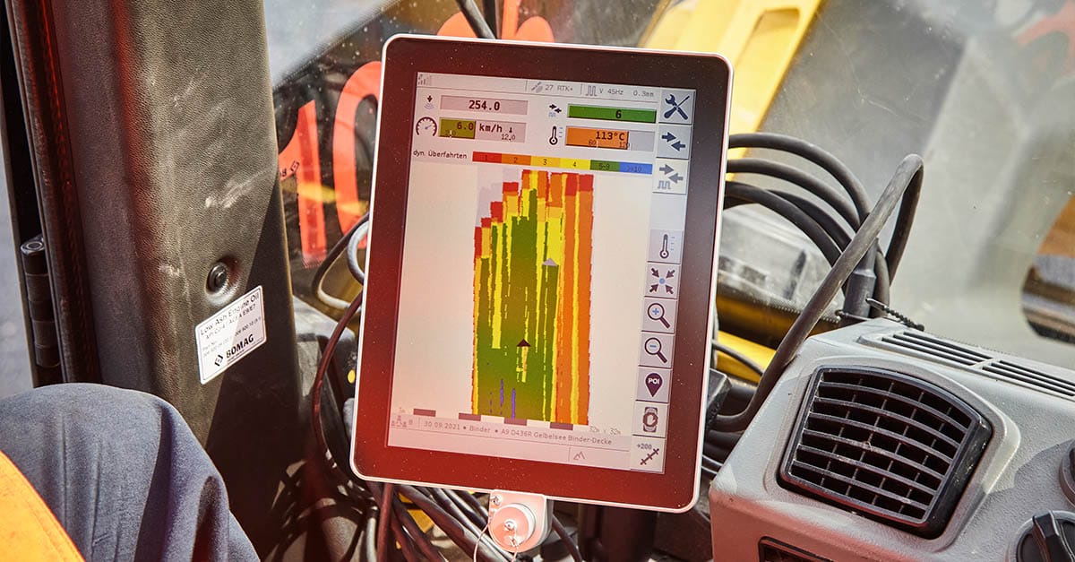A close-up of a tablet displaying a color-coded bar graph with labels and statistics in a construction vehicle. The piece of technology is mounted near the vehicle's dashboard, with black cables connected to it. There are reflections on the screen indicating sunlight.