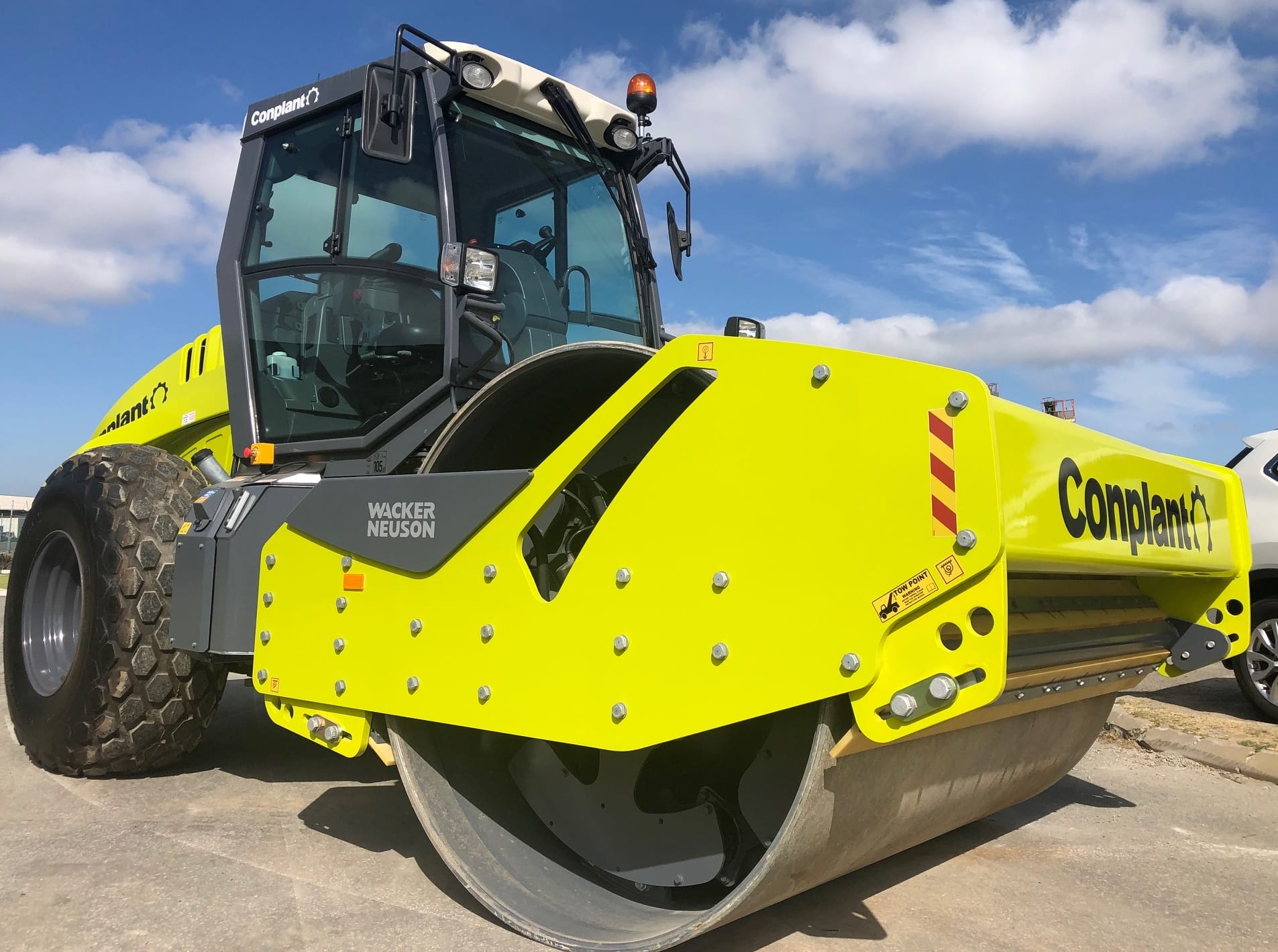 A bright yellow road roller with the "Conplant" logo and "Wacker Neuson" label on its side is parked on a paved area under a clear blue sky. The machine, an impressive example among soil compactors, has large tires and a massive front roller designed for compacting surfaces.