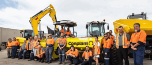 A group of Conplant construction workers in orange high-visibility shirts stand and sit in front of a range of machinery, including an excavator, a Wacker Neuson telehandler, and a dump truck. They appear to be posing for a group photo at the construction site.