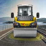 A vibrating roller machine featuring advanced Oscillation Technology rolls over a bridge with scenic water and hills in the background. The yellow machine, accented in black, proudly displays the logo "W" on its front. Flanked by railings on both sides, this setup ensures Quality Compaction throughout.