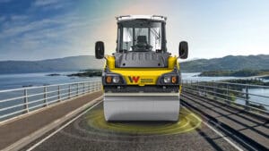 A vibrating roller machine featuring advanced Oscillation Technology rolls over a bridge with scenic water and hills in the background. The yellow machine, accented in black, proudly displays the logo "W" on its front. Flanked by railings on both sides, this setup ensures Quality Compaction throughout.