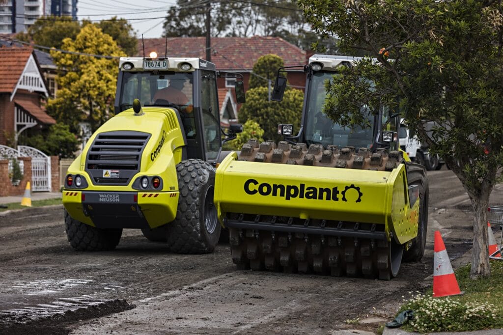 Two Conplant road rollers, one with a smooth drum and the other with a padded drum, are working on a residential street resurfacing project. The street is partialy closed off with traffic cones, and houses with gardens are visible in the background.