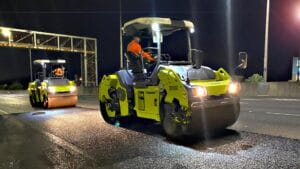 Two workers use asphalt resurfacing equipment, paving at night under bright lights.