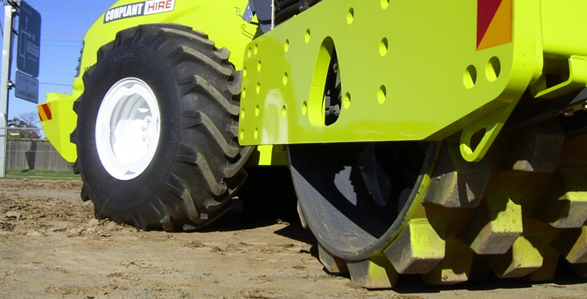 Close-up of a bright yellow construction vehicle compacting clay soil on a dirt surface.