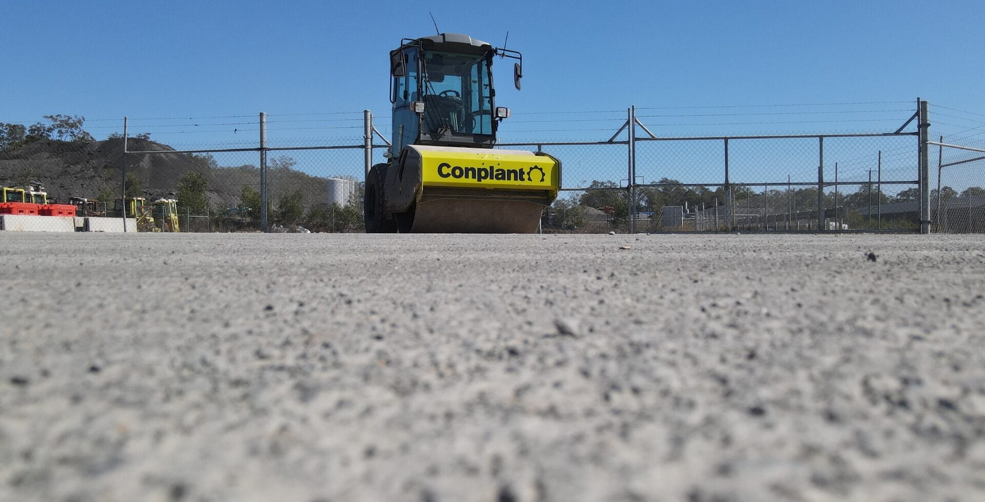 A road roller for soil compaction on gravel, seen from a low angle with fence and blue sky behind.