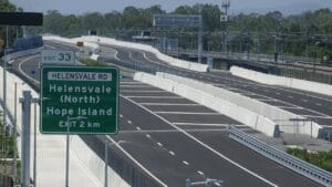 Green highway exit sign for Helensvale Rd, Helensvale (North) and Hope Island, exit in 2 km, on a multi-lane road with concrete barriers.