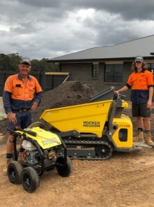 Two construction workers, Brady and Wes Faulkner, in orange safety shirts and work boots stand beside a yellow Wacker Neuson machine and a portable generator on a dirt site. A house with a dark roof is in the background, under a cloudy sky.