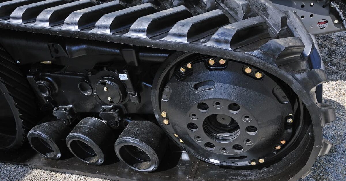 Close-up image of the track and wheel assembly of a heavy-duty vehicle, such as a tank or construction equipment. The rubber excavator tracks feature durable rubber with metal components, and three smaller wheels are visible, aiding in mobility over rough terrain.