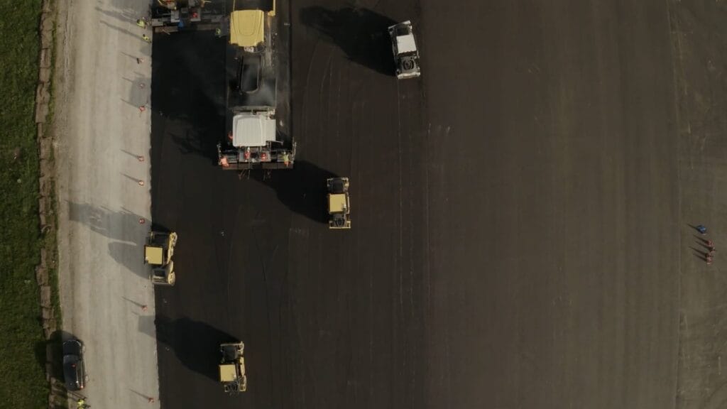 Aerial view of construction vehicles working on a large, dark asphalt surface for the runway overhaul at Salzburg Airport. Several machines, like a Völkel road roller and a paver, are spread out on the site. Two small figures stand near the edge on the right, bordered by grass and gravel.