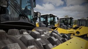 Close-up of the spiked roller on a large yellow compactor, a type of padfoot roller, with similar vehicles in the backdrop. The partly cloudy sky adds depth to this industrial scene, highlighting the benefits and applications these machines offer in construction.