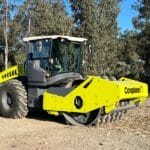 A large yellow road roller with a protective cab, akin to padfoot rollers, is parked on a dirt path. The machine's wide, heavy-duty wheels are perfect for applications like compacting soil and gravel. Trees and a small fence are in the background under a clear sky.