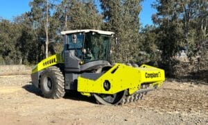 A large yellow road roller with a protective cab, akin to padfoot rollers, is parked on a dirt path. The machine's wide, heavy-duty wheels are perfect for applications like compacting soil and gravel. Trees and a small fence are in the background under a clear sky.