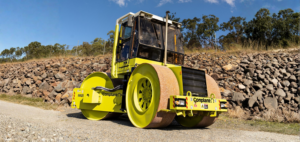 Yellow road roller with twin front rollers on a gravel surface beside a stone embankment and trees under a blue sky.