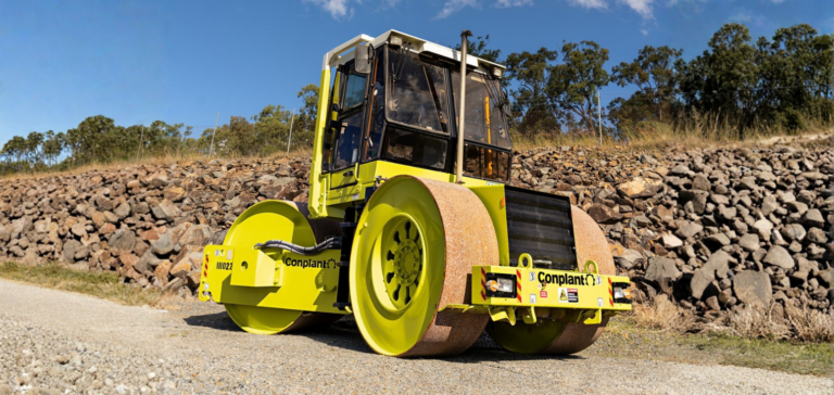 Yellow road roller with twin front rollers on a gravel surface beside a stone embankment and trees under a blue sky.