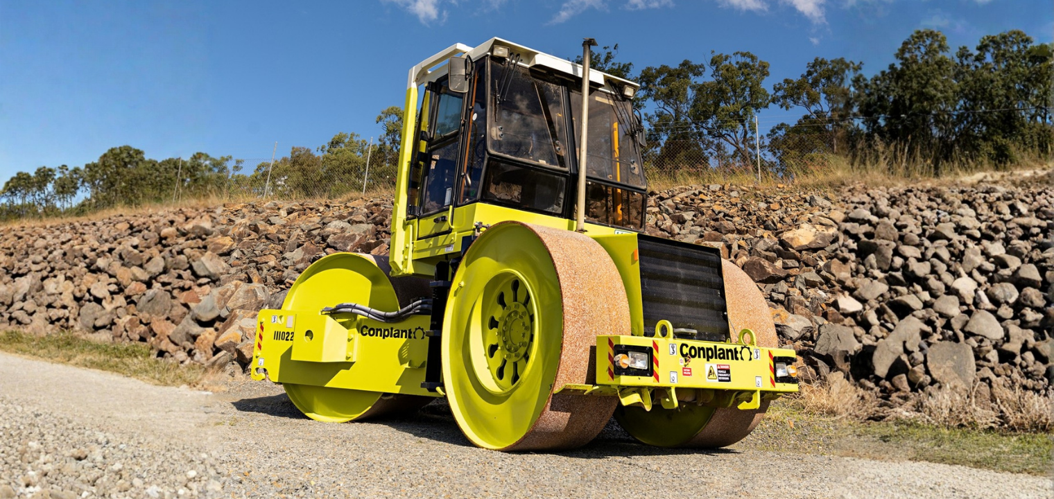 proof rolling blog - Conplant Pty Ltd - Conplant Pty Ltd Yellow road roller with twin front rollers on a gravel surface beside a stone embankment and trees under a blue sky.
