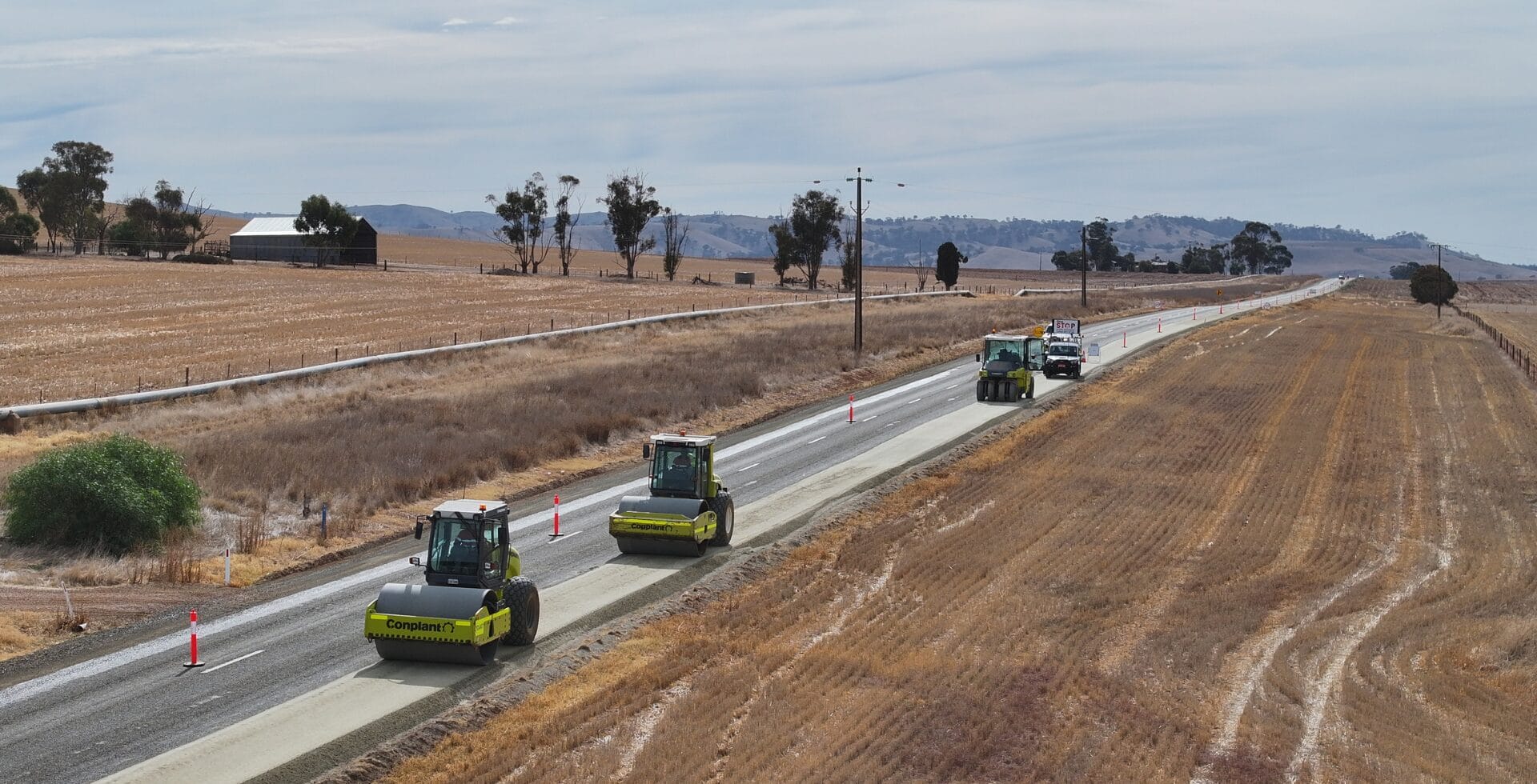 Three road rollers and trucks reduce rework on a rural road construction site between dry fields.