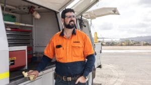 Man in orange work shirt stands by utility truck; regular inspections ensure equipment lifespan.