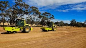 Two yellow construction rollers are diligently working on a dirt road under a blue sky with scattered clouds. Their effort in compaction promises smooth finishes. Trees line the background, partially shading the cleared landscape.