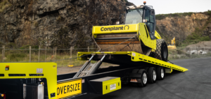 Yellow road roller on a low-boy trailer marked OVERSIZE, secured with chains, ready for transport at a construction site near rocky hillsides.