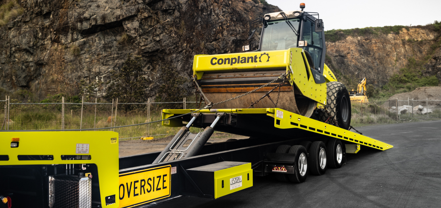 Yellow road roller on a low-boy trailer marked OVERSIZE, secured with chains, ready for transport at a construction site near rocky hillsides.