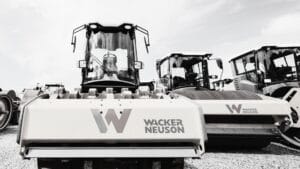 A row of Wacker Neuson construction vehicles, including steamrollers used for soil compaction, is parked on a gravel lot. The large company logo is visible on the nearest machine in this black-and-white image.