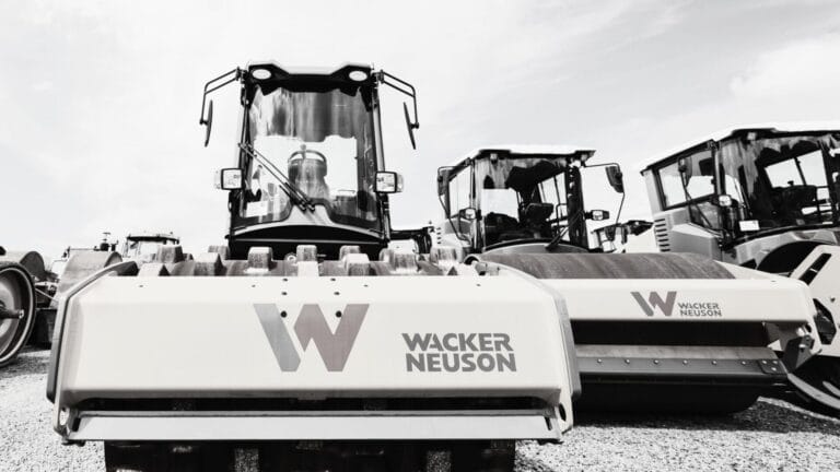 A row of Wacker Neuson construction vehicles, including steamrollers used for soil compaction, is parked on a gravel lot. The large company logo is visible on the nearest machine in this black-and-white image.