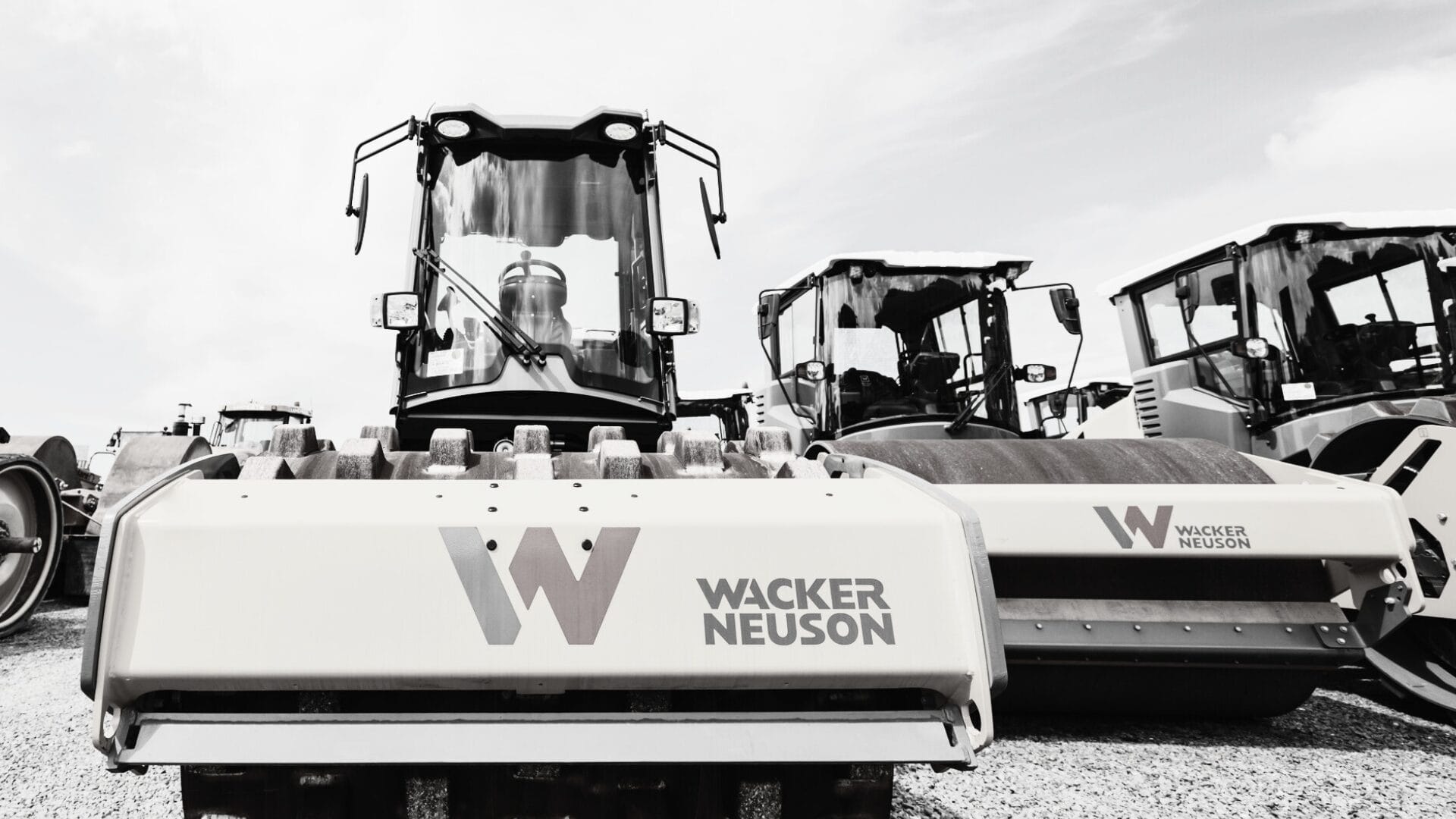 A row of Wacker Neuson construction vehicles, including steamrollers used for soil compaction, is parked on a gravel lot. The large company logo is visible on the nearest machine in this black-and-white image.