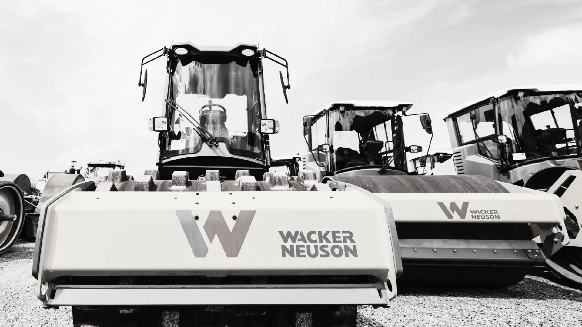 A row of Wacker Neuson construction vehicles, including steamrollers used for soil compaction, is parked on a gravel lot. The large company logo is visible on the nearest machine in this black-and-white image.